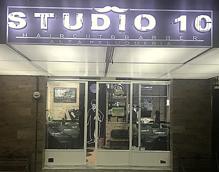 Fachada de Studio 10 de noche: letrero iluminado STUDIO 10 Haircut and Barber, Alta peluquería, con un bigote encima; detrás del vidrio, los sillones con capa negra y la silueta de bombín y bastón en la pared