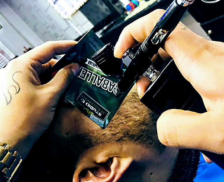 Manos del barbero perfilando la barba de un cliente con aerógrafo y una plantilla con el logo de Studio 10
