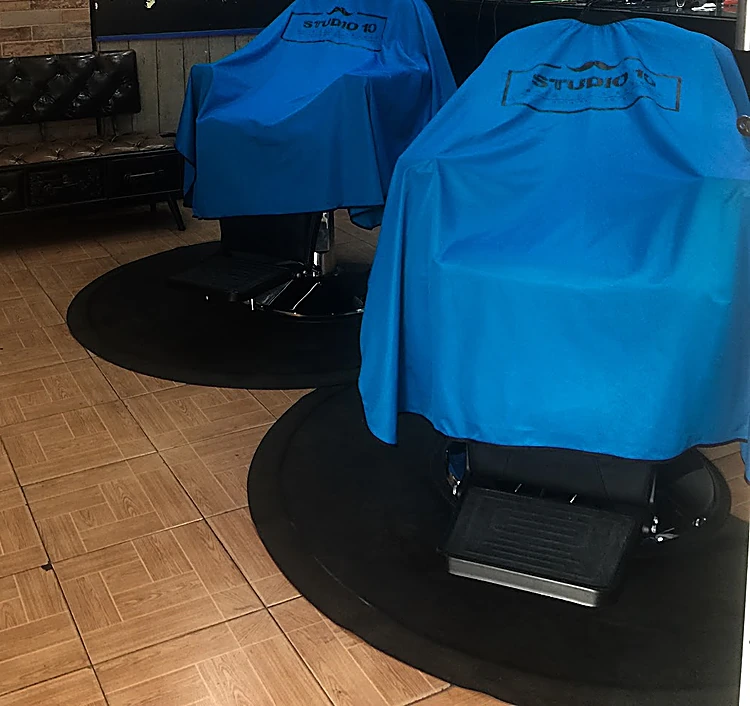 Dos sillones de barbero cubiertos con capas azul eléctrico con el logo de Studio 10, sobre tapetes redondos negros y piso de loseta